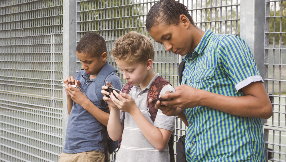 Three children lean against a chain link fence staring and typing on their smart phones