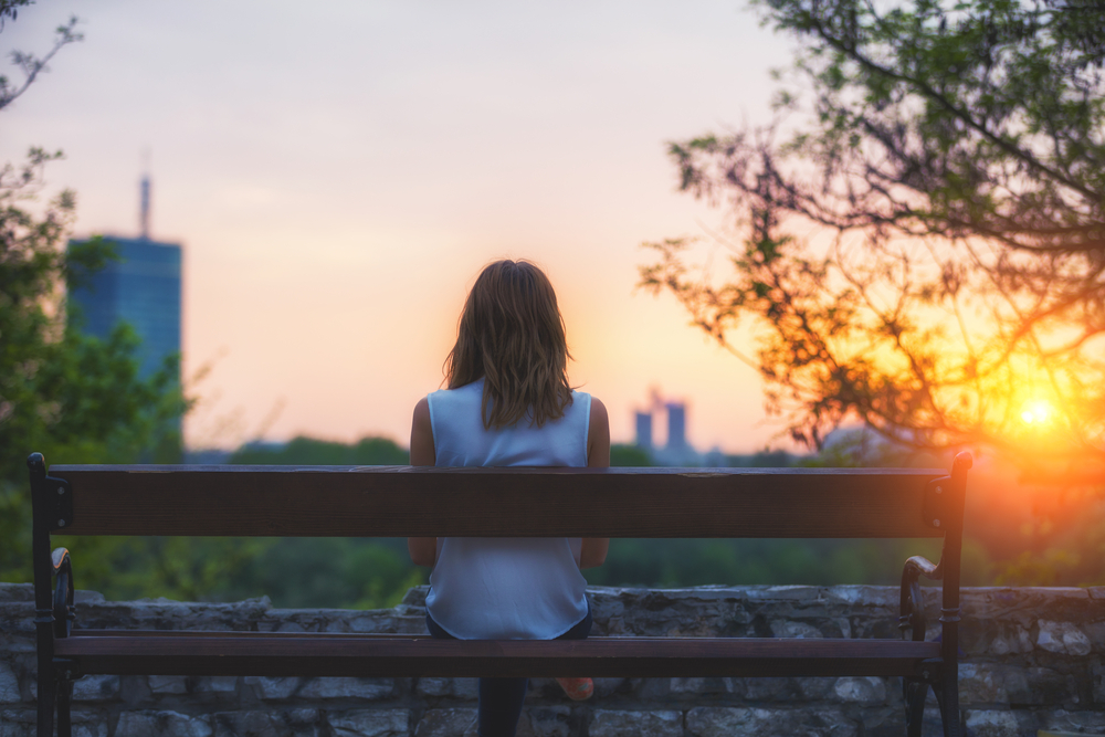 A young woman sits on a park bench at sunset struggling with depression and lack of family assistance
