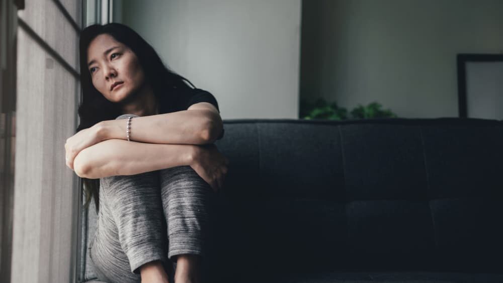 A forlorn woman hugging her knees to her chest as she listlessly stares out a window.