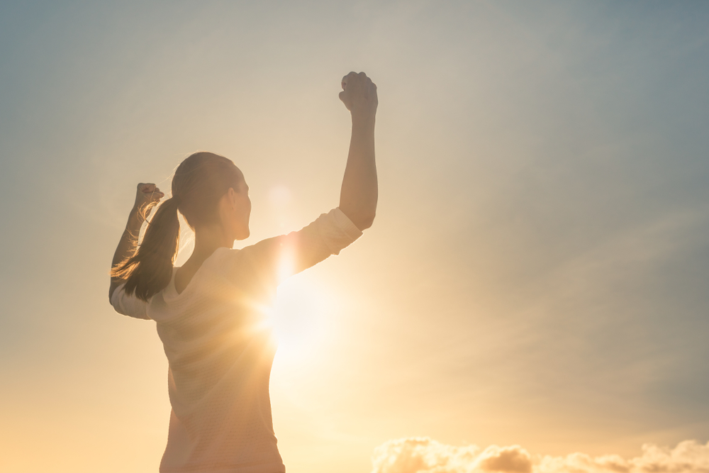 A healthy young woman celebrates completing a hike as the sun is setting as part of her mental health goal setting plan